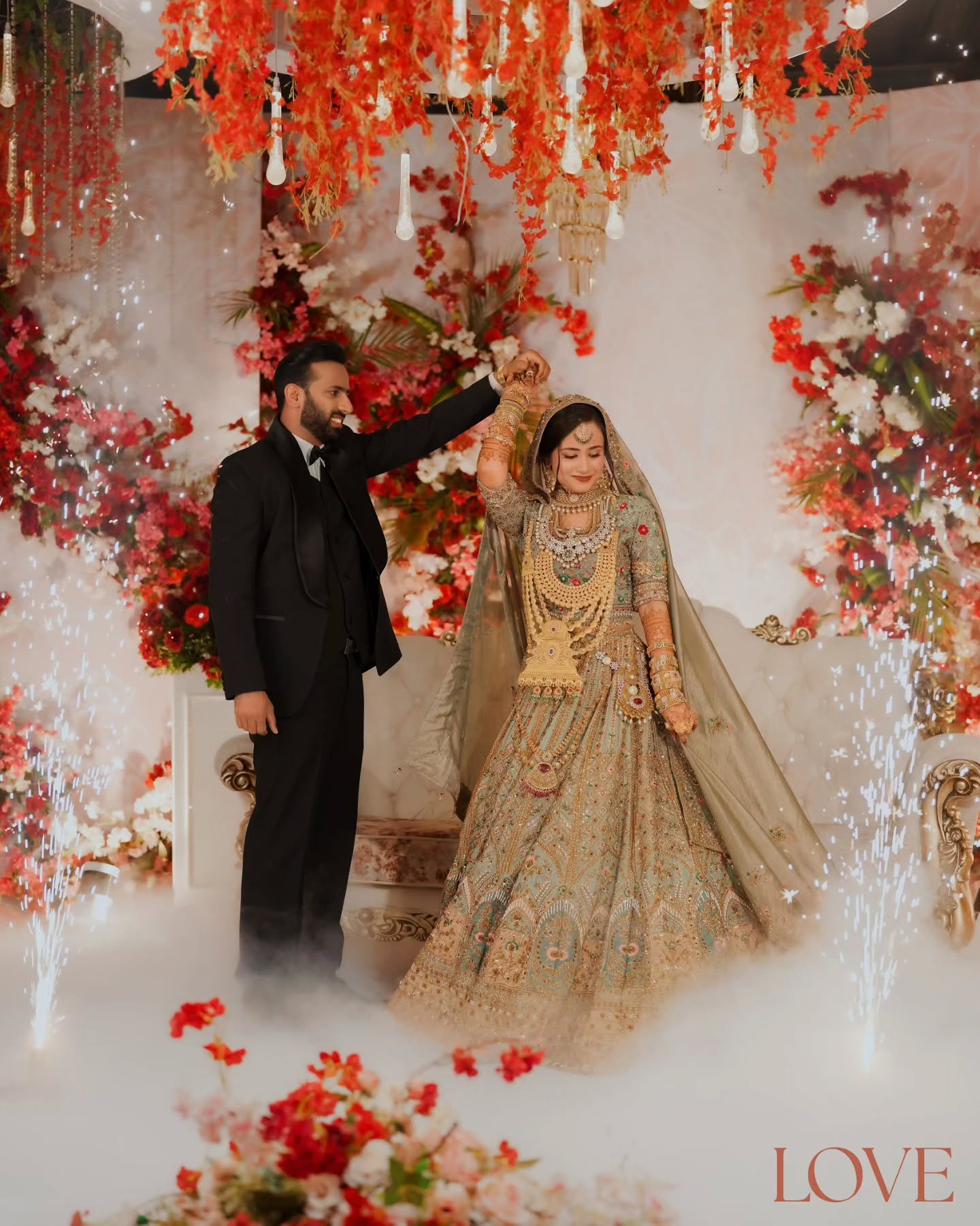 Bride and groom twirling beneath a red floral ceiling amid drifting mist and sparklers