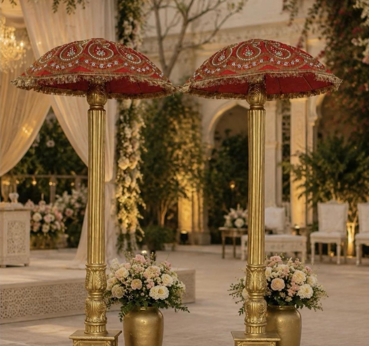 Two gold pillars topped with crimson tasselled umbrellas beside urns of blush roses